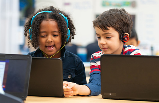 Two elementary school students wearing headphones and working on laptops